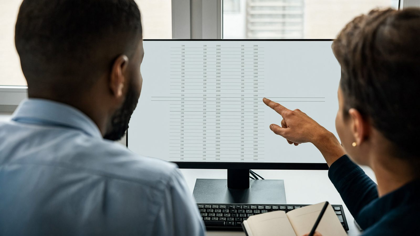 Two colleagues at a monitor reviewing a long customer list, one pointing at a row on screen.