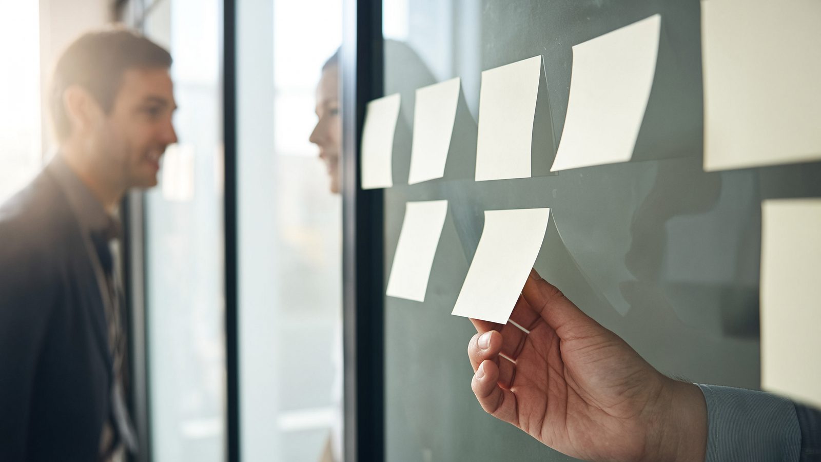 Hands moving sticky notes between columns on a glass wall, a colleague watching.