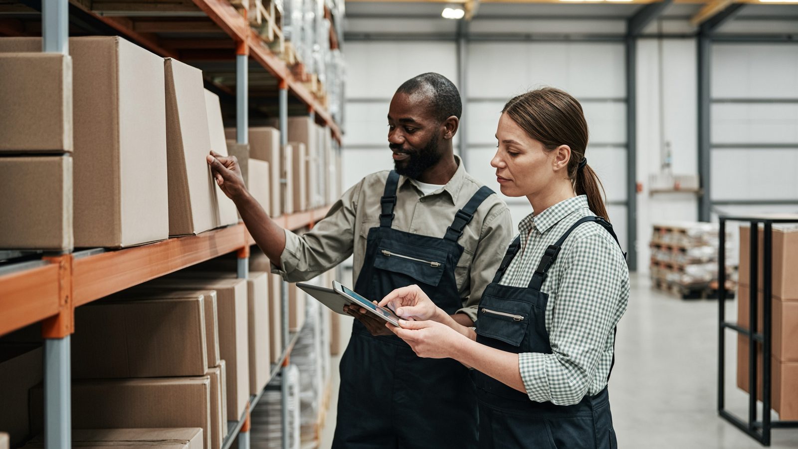 Two warehouse staff counting stock on a rack, one holding a tablet.